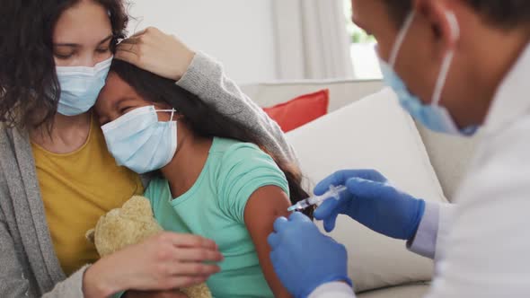 Hispanic male doctor making injection to girl with mother at home, all wearing face masks,