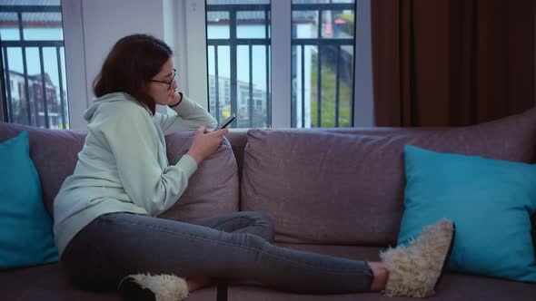 Girl with Glasses Looks Out of a Window and Texting on Her Mobile Phone While Sitting on the Couch