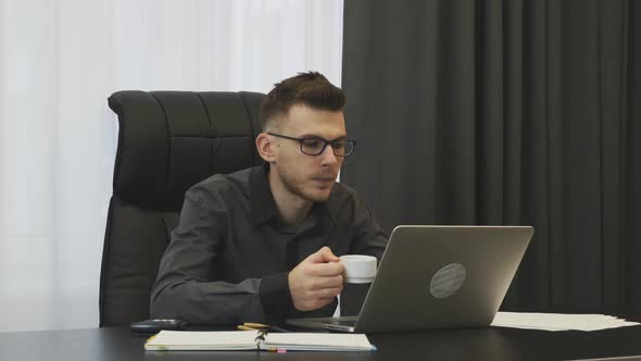 Young businessman in suit and glasses drinks coffee in his modern office. Business corporate concept