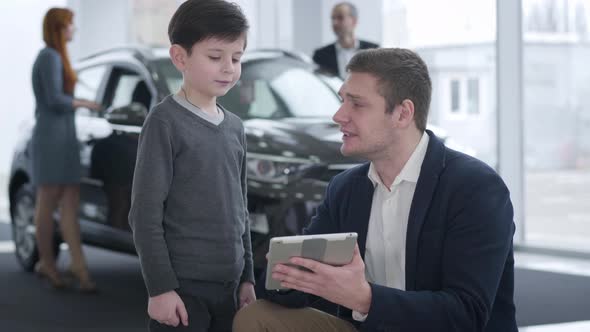 Portrait of Positive Young Caucasian Car Dealer Entertaining Little Brunette Boy While His Parents