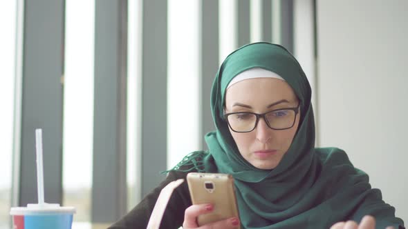 Young Muslim Woman in a Hijab Is Sitting at a Table By the Window Using the Phone and Eating