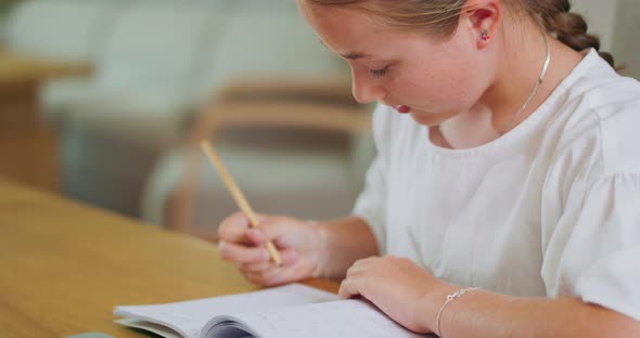 Closed Focused Teen Girl at the Desk Does Tasks in Workbook and Checks Them on Laptop Background is alt