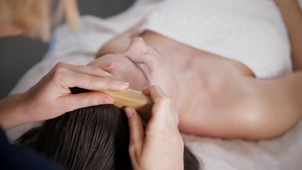 Massage Session  Young Woman Therapist Using Wooden Piece on the Face of Her Client