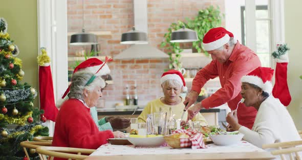 Caucasian senior man in santa hat carving turkey at christmas dinner table with diverse friends
