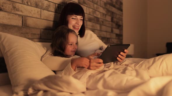 Mother and Daughter Using Tablet Pc Lying in Bed Under the Blanket and Spending Their Evening Time