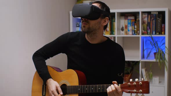 A man in a virtual reality helmet is actively playing an acoustic guitar sitting in a room