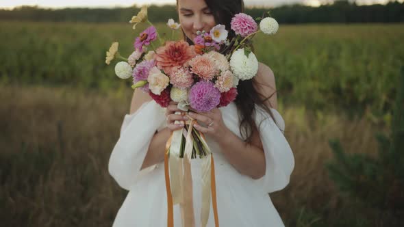 A Beautiful Smiling Bride Stands By the Field and Brings a Delicate Bouquet of Dahlias with Ribbons