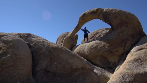 A young man trail running with his dog past a natural rock arch in a mountainous desert.