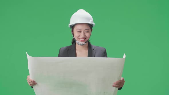 Smiling Asian Female Engineer With Safety Helmet Looking At Blueprint In The Green Screen Studio