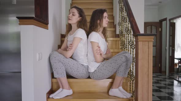 Dissatisfied Argued Twin Sisters in Pajamas Sitting on Stairs Back to Back