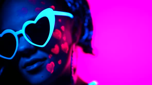 Portrait of fashion model woman with heart shaped glasses in neon light. Fluorescent unusual makeup