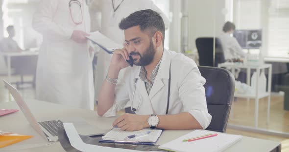 Male Doctor Talking on Phone with Patient and Working at Laptop