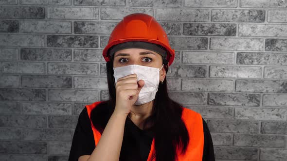 Female Construction Worker in Overalls and Medical Mask Coughing on Background of Gray Brick Wall