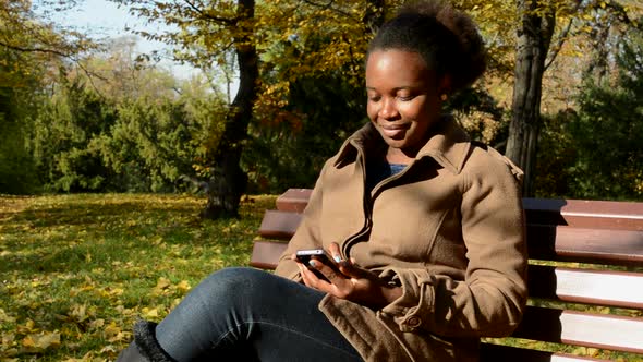 Young Beautiful African Happy Girl Sits on Bench in Woods, Works on Phone and Observes Landscape