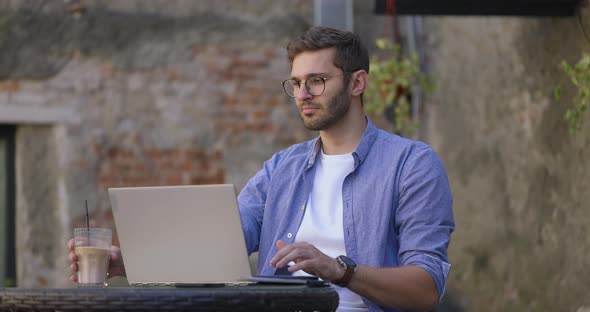 Men Wearing Eyeglasses and Blue Shirt Working on Laptop and Drink Ice Coffee in Outdoor Cafe