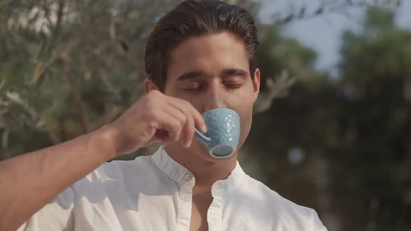 Attractive young fashion man in a suit drinks coffee in the morning outdoors in the garden