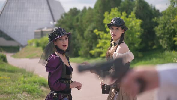 Scared Beautiful Ladies in Steampunk Outfit Looking at Blurred Gun in Hand at Front with Frightened