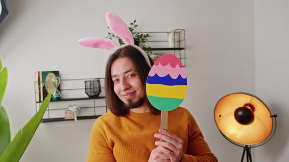 Smiley Bearded Caucasian Man Hiding Behind Colorful Easter Egg Cardboard and Wearing Bunny Ears