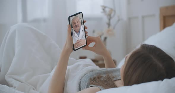 Female Medical Assistant Wears White Coat, Headset Video Calling Distant Patient on Smartphone