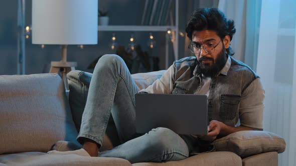 Freelancer Guy Arab Indian Man Bearded Male User Wearing Glasses Sitting on Sofa at Night Late Time