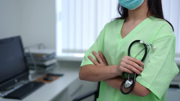 Unrecognizable Female Doctor in Coronavirus Face Mask with Crossed Hands and Stethoscope Standing in