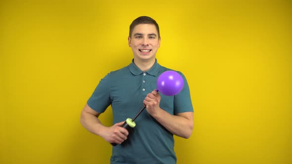 A Young Man Inflates a Purple Balloon with a Pump on a Yellow Background. Man in a Green Polo.