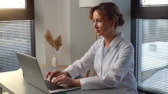 Serious Woman Doctor in White Coat Working Typing on Laptop Computer Looking on Display Screen