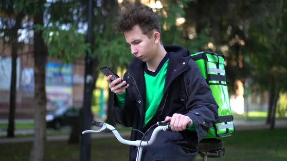 Delivery Man with Green Bagpack and a Bicycle Is Looking for the Client's Address in the Phone