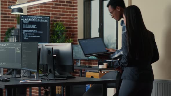 Two Software Developers Holding Laptop with Coding Interface Walking Towards Desk and Sitting Down