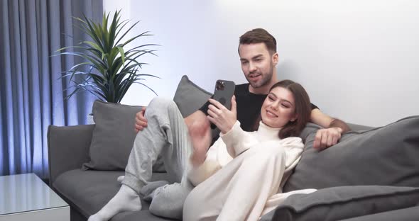 Young Couple Takes a Selfie on a Smartphone a Guy and a Girl Sit on the Sofa in the Living Room and