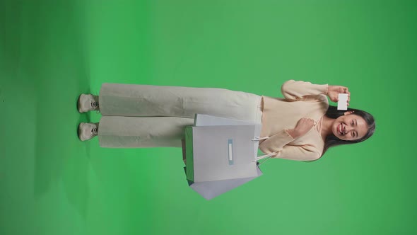 Full View Of An Asian Shopping Woman With Shopping Bags Holding A White Card On Green Screen