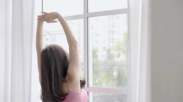 Beautiful young asian woman wake up and walking to window with open curtains in the morning.