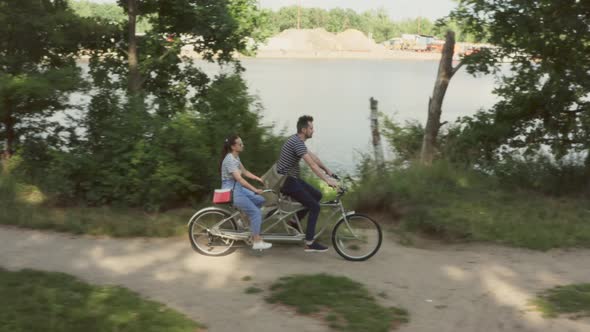 A young couple rides a tandem two-seater bike along the riverbank