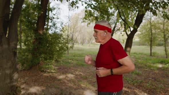 Male Senior Person Running Along the Road in Park. Mature Runner Man Training, Listening Music