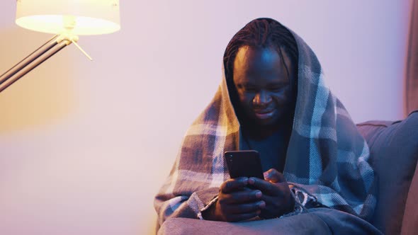 Young African American Black Man Covered with Blanket Over the Head Using Smartphone with Smile
