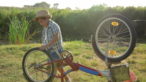 Curious Kid Is Spinning Bicycle Wheel and Pedals at Rural on Background Nature in Backlight