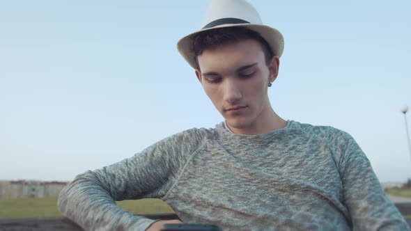Young Attractive Guy in a Hat Reads Messages on His Phone and Smiles