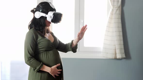 Young pregnant female in VR goggles standing near window