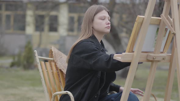 Concentrated Woman Painter Sitting in Front of Wooden Easel Drawing a Picture