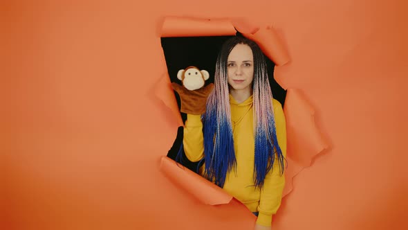Young Woman with Soft Puppet Toy on Hand Looking Out of Hole of Orange Background