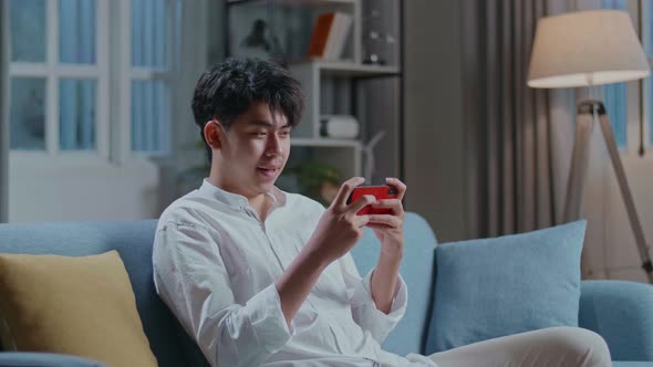 Asian Man Playing Game On Smartphone And Smiling While Sitting On Sofa In The Living Room