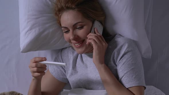 Smiling Woman in Bed Holding Pregnancy Test and Talking Phone