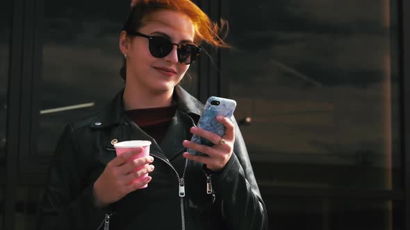 Young Woman Drinking Coffee on the Street, Typing Texting a Mobile Phone, While Walking in an Urban