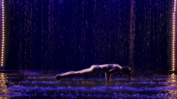 Beautiful Young Woman Stands in the Kumbhakasana Pose with Studio Blue Light and Water Rain. Female