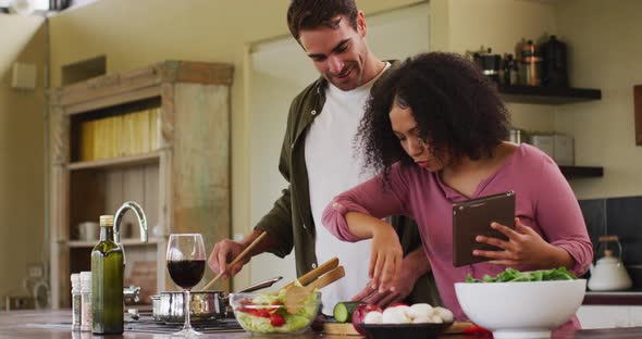 Happy diverse couple preparing a meal together in kitchen, using recipe on tablet