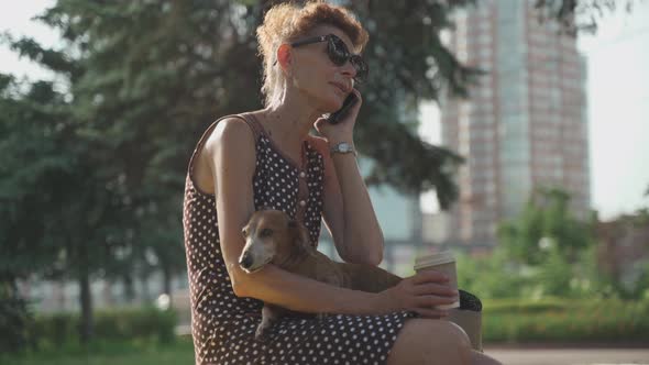 An Elderly Woman Talking on the Phone Holding a Small Dachshund Dog and Drinking Coffee to Go While