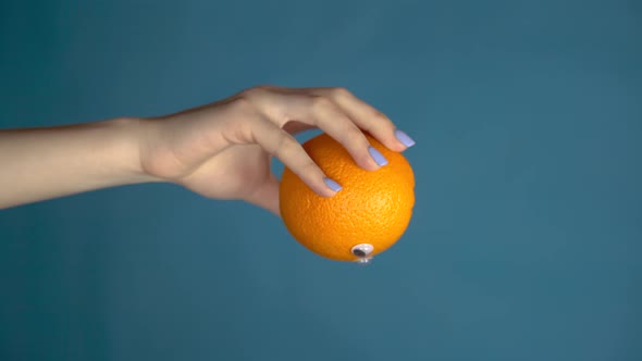 Orange with Eyes in a Woman Hand Close Up. Orange Looks Around on a Blue Background. Vertical Video.