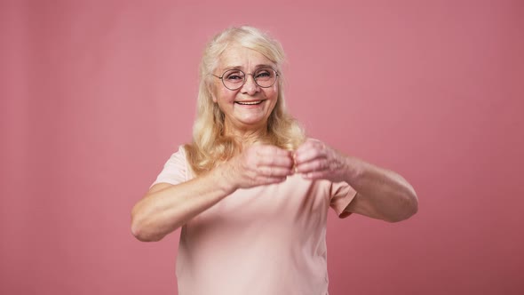 Positive Senior Woman in Glasses Showing Heart Gesture Near Her Chest Expressing Kindness Pink