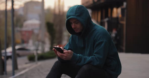 Man in Hoodie Using Smartphone Sitting in the City