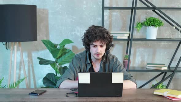 Young Curly Haired Man Using Laptop Computer with Headset to Talking Online